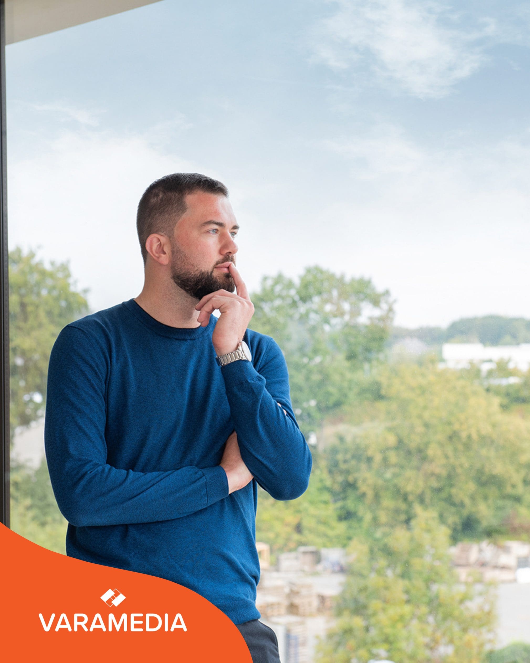 Man in blauwe trui kijkt nadenkend uit raam met stadsgezicht