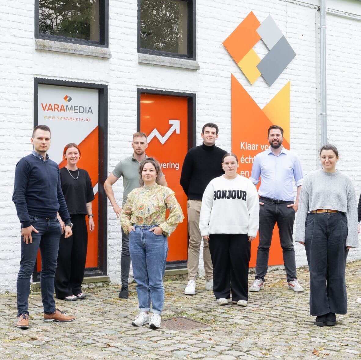 Teamfoto van het Varamedia marketingbureau met medewerkers buiten het kantoor en Varamedia-branding op de achtergrond | Varamedia
